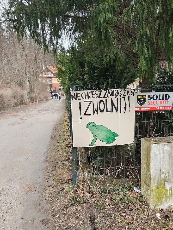 Na ogrodzeniu przy piaszczystej drodze wisi ręcznie wykonana tabliczka z rysunkiem zielonej żaby i prośbą o zwolnienie prędkości, by nie zabijać płazów. Tabliczka umieszczona jest obok profesjonalnej tablicy informacyjnej o ochronie obiektu, co dodatkowo przyciąga wzrok przechodniów.