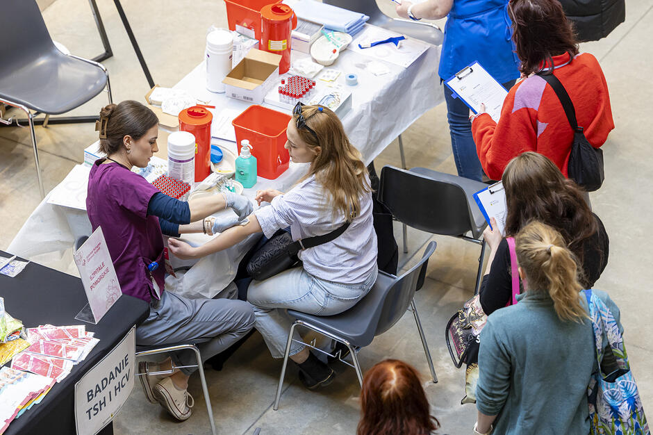 Grupa ludzi zebranych wokół stołu w pokoju. Na zdjęciu cztery osoby, trzy kobiety i jeden mężczyzna. Stół jest pokryty białym obrusem i znajdują się na nim różne przedmioty, w tym butelki, pudełka i inne artykuły medyczne. Kobieta po lewej stronie nosi purpurową koszulę i wydaje się być pielęgniarką lub lekarzem. Siedzi na krześle i ma w ręku schowek i długopis. Obok niej jest inna kobieta w pomarańczowej kurtce i noszący stetoskop wokół szyi. Po prawej stronie również trzyma schowek. Przed nimi można zobaczyć kilka innych osób siedzących na ławkach i przyglądających się scenie.