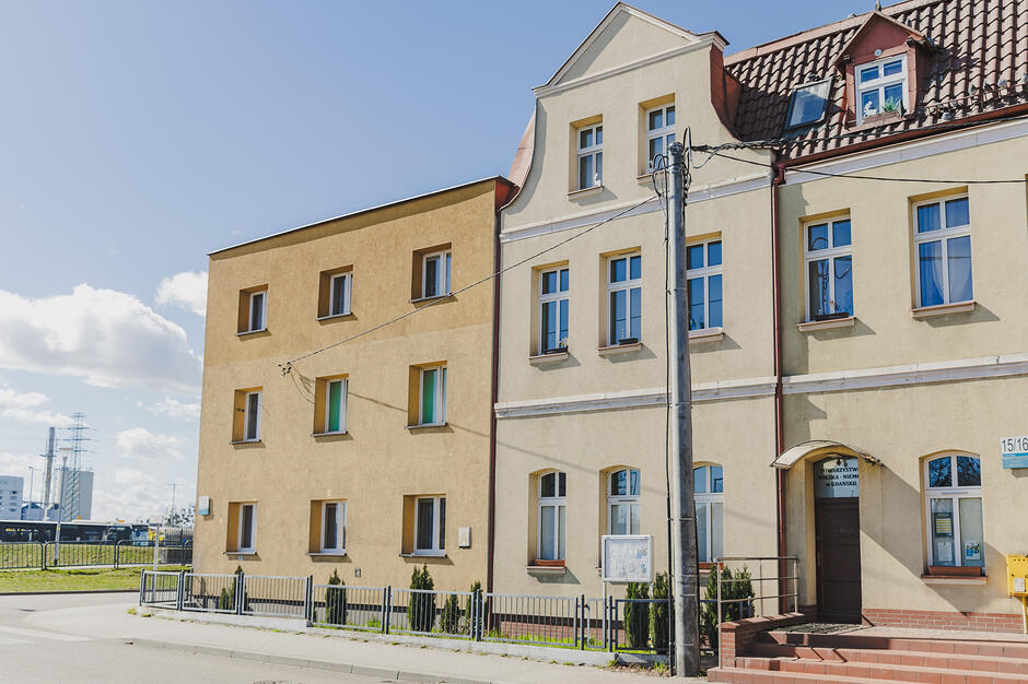 Zdjęcie przedstawia dwa przylegające do siebie wielopiętrowe budynki o jasnych, żółtawych elewacjach, z których jeden posiada ozdobny szczyt i spadzisty dach. Przed obiektami znajduje się niskie ogrodzenie, chodnik oraz słup energetyczny, a w tle widać infrastrukturę przemysłową pod jasnym niebem.