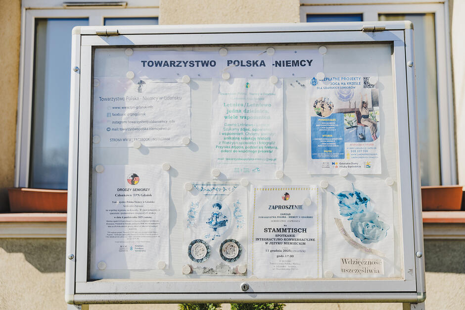 Zdjęcie przedstawia oszkloną gablotę informacyjną Towarzystwa Polska-Niemcy, w której umieszczono różne ogłoszenia, zaproszenia i plakaty. Wewnątrz znajdują się między innymi informacje o spotkaniach językowych „Stammtisch”, zajęciach jogi dla seniorów oraz dane kontaktowe stowarzyszenia.