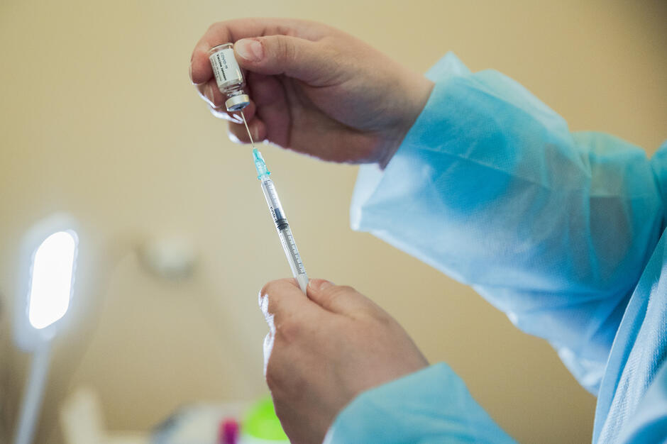 na zdjęciu dwie dłonie w lekarskim fartuchu ochronnym, trzymają szczepionkę