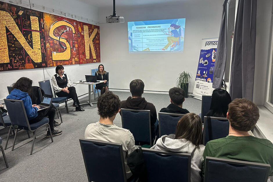 grupa młodzieży siedzi na krzesłach w sali warsztatowej, widać ich od tyłu. Przed nimi siedzą dwie kobiety. Za nimi ekran z prezentacją.