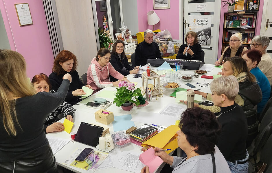 Kilkanaście osób siedzi wokół stołu w sali podczas warsztatu. Jedna kobieta stoi i pokazuje coś pozostałym ręką.