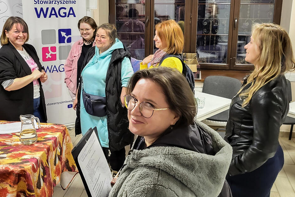 Kilka kobiet stoi w sali podczas warsztatów, uśmiechają się, kobieta na pierwszym planie stoi bokiem i obraca twarz do aparatu.