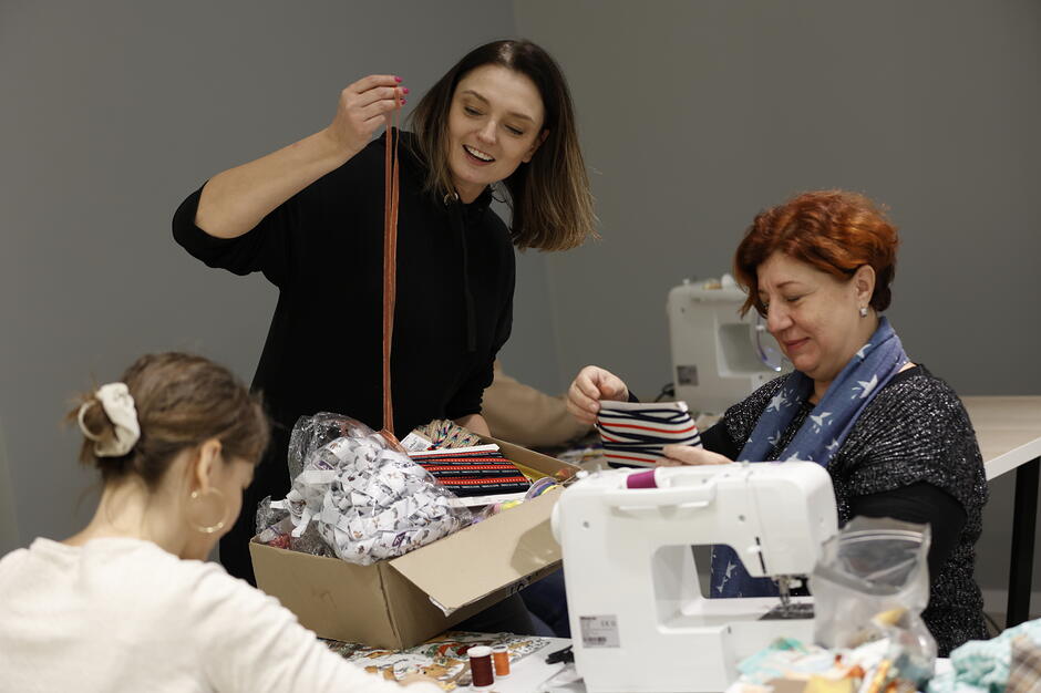 Trzy kobiety przy stole i maszynie do szycia. Dwie z nich siedzą, jedna stoi i śmieje się.