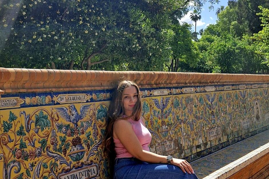 na zdjęciu kobieta siedząca na długiej zdobionej ptakami i kwiatami ławie, w tle roślinność.