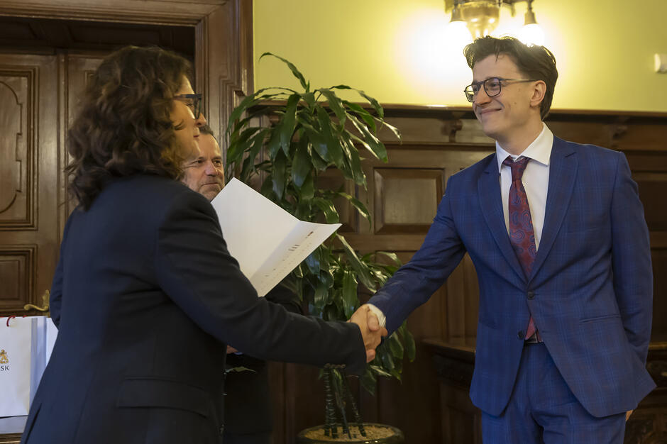 Kobieta i mężczyzna w eleganckich strojach podają sobie dłonie w oficjalnym geście gratulacji lub powitania. W tle widać roślinę doniczkową, drewniane panele ścienne oraz fragment białej torby z logo, co sugeruje formalne spotkanie lub ceremonię.