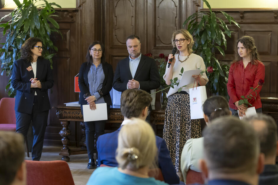 Grupa osób stoi przed audytorium w zabytkowym wnętrzu, gdzie jedna z kobiet przemawia do mikrofonu, trzymając różę i białą torbę upominkową. Pozostali uczestnicy, ubrani w stroje wizytowe, przysłuchują się wystąpieniu na tle ozdobnych, drewnianych paneli i roślin doniczkowych.