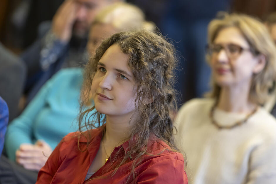 Młoda kobieta o kręconych włosach, ubrana w czerwoną koszulę, patrzy z uwagą przed siebie podczas oficjalnego zgromadzenia. W rozmytym tle widoczne są inne osoby, co sugeruje uczestnictwo w wykładzie, konferencji lub podobnym wydarzeniu publicznym.