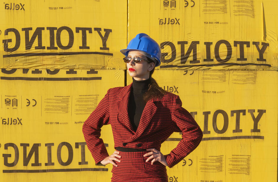 na zdjęciu kobieta w niebieskim kasku roboczym, na tle żółtej ściany z odwróconymi napisami GNIOTY