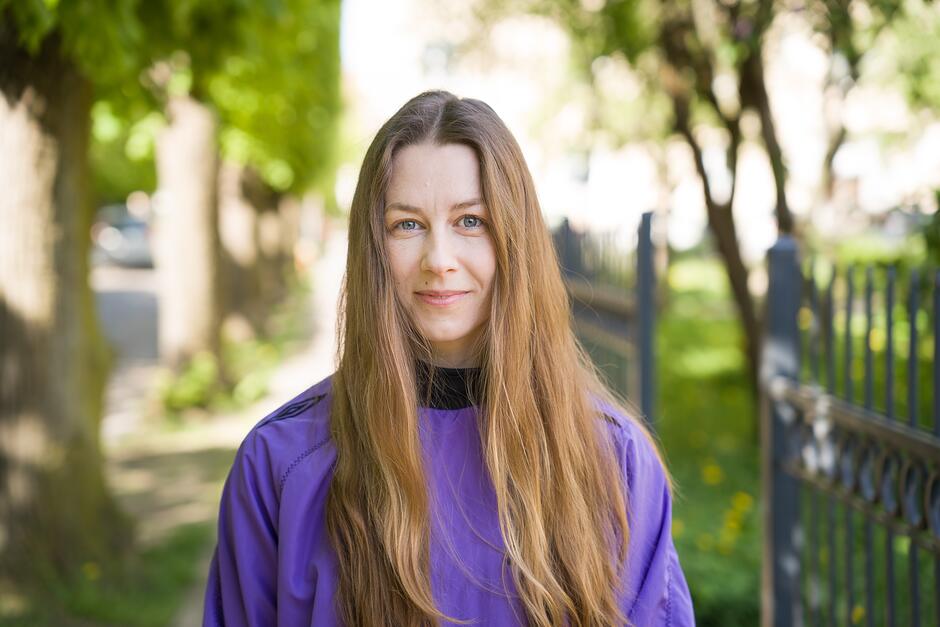 Na zdjęciu kobieta z włosami rozpuszczonymi, sięgającymi za ramiona, w tle zieleń