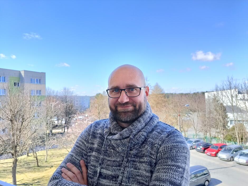 
mężczyzna stojący na balkonie, widoczny od pasa w górę, z założonymi rękami. Nosi okulary i gruby, szary sweter z wysokim kołnierzem. W tle drzewa bez liści, zaparkowane samochody oraz budynki mieszkalne. Zdjęcie wykonane w ciągu dnia, przy jasnym niebie.
