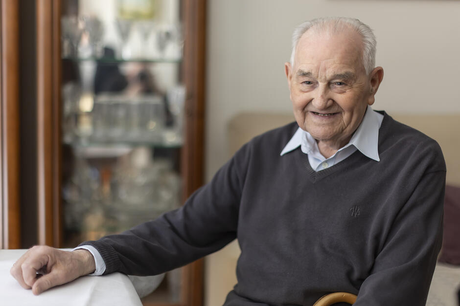 Starszy mężczyzna pozuje do zdjęcia w mieszkaniu siedząc za stołem, uśmiecha się.