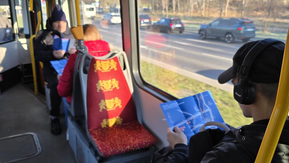 Ludzie jadą tramwajem, widzimy trzy siedzące osoby, dwie z nich czytają ulotki