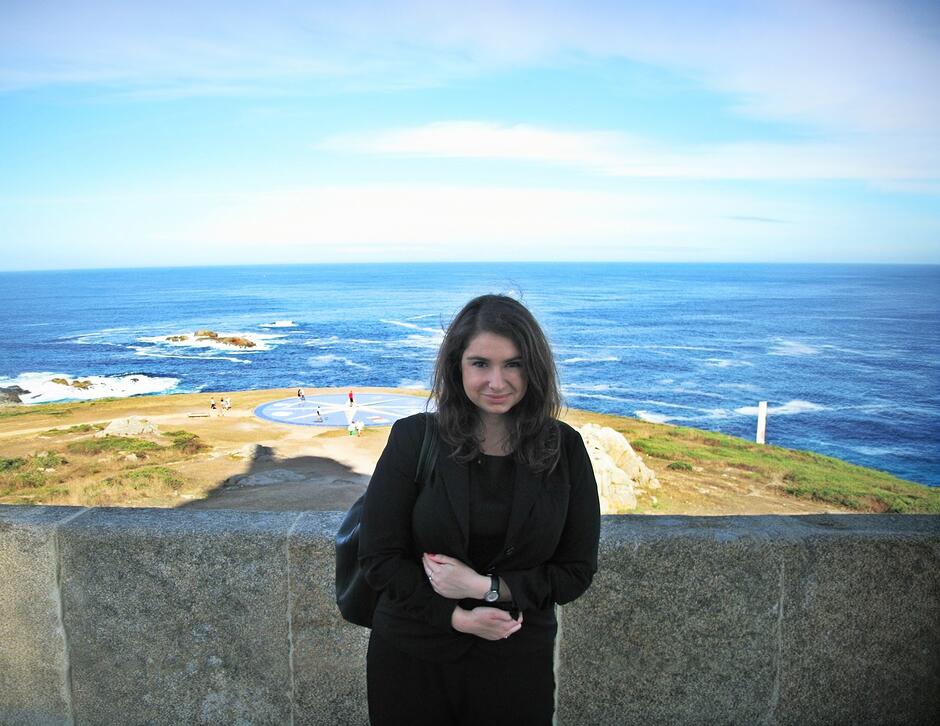 na zdjęciu kobieta o ciemnych włosach za ramiona, ubrana w czarny płaszcz, w tle krajobraz morskich fal