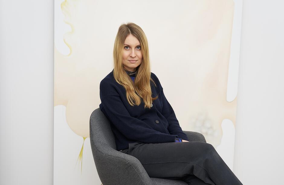 na zdjęciu kobieta o jasnych włosach, w ciemnym ubraniu, siedząca w fotelu, na tle namalowanego obrazu w ciepłych odcieniach.