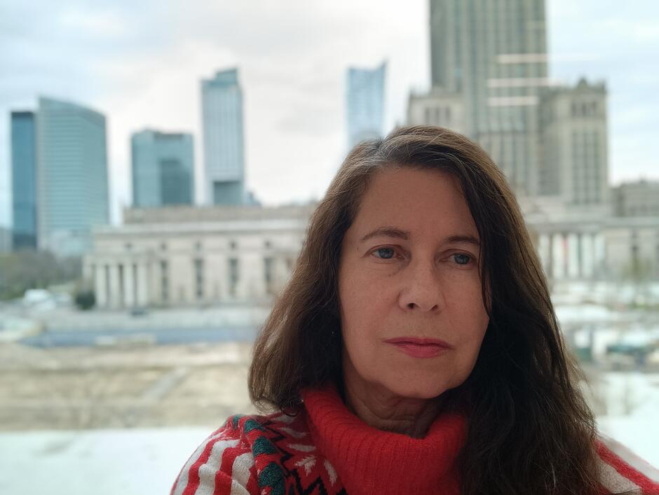 Na zdjęciu kobieta z rozpuszczonymi ciemnymi włosami, patrząca na wprost, za nią budynki nowoczesnego miasta.