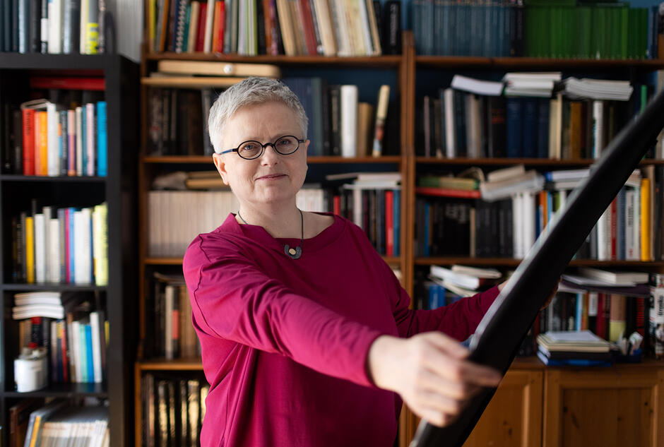 Na zdjęciu kobieta z krótkimi włosami, w okularach, na tle półek z książkami, w ręce trzyma drewniany obiekt.