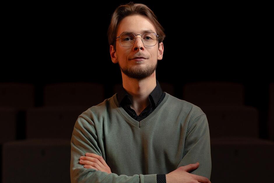 na zdjęciu mężczyzna patrzący przed siebie, w okularach, z lekkim zarostem, ma skrzyżowane przed sobą ręce