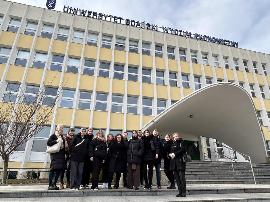 Zdjęcie przedstawia grupę ludzi pozujących przed budynkiem Wydziału Ekonomicznego Uniwersytetu Gdańskiego w Sopocie. Nad wejściem, które wyróżnia się charakterystycznym, szerokim zadaszeniem w kształcie łuku, widnieje duży napis z nazwą uczelni i wydziału.