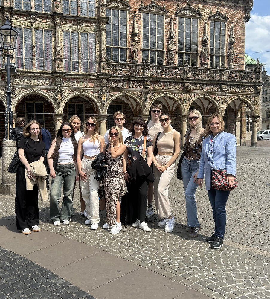 Grupa młodych ludzi pozuje do wspólnego zdjęcia na brukowanym placu przed zabytkowym, bogato zdobionym budynkiem z arkadami. Uczestnicy są ubrani w swobodne, letnie stroje, a towarzyszy im uśmiechnięta kobieta w niebieskiej marynarce. 