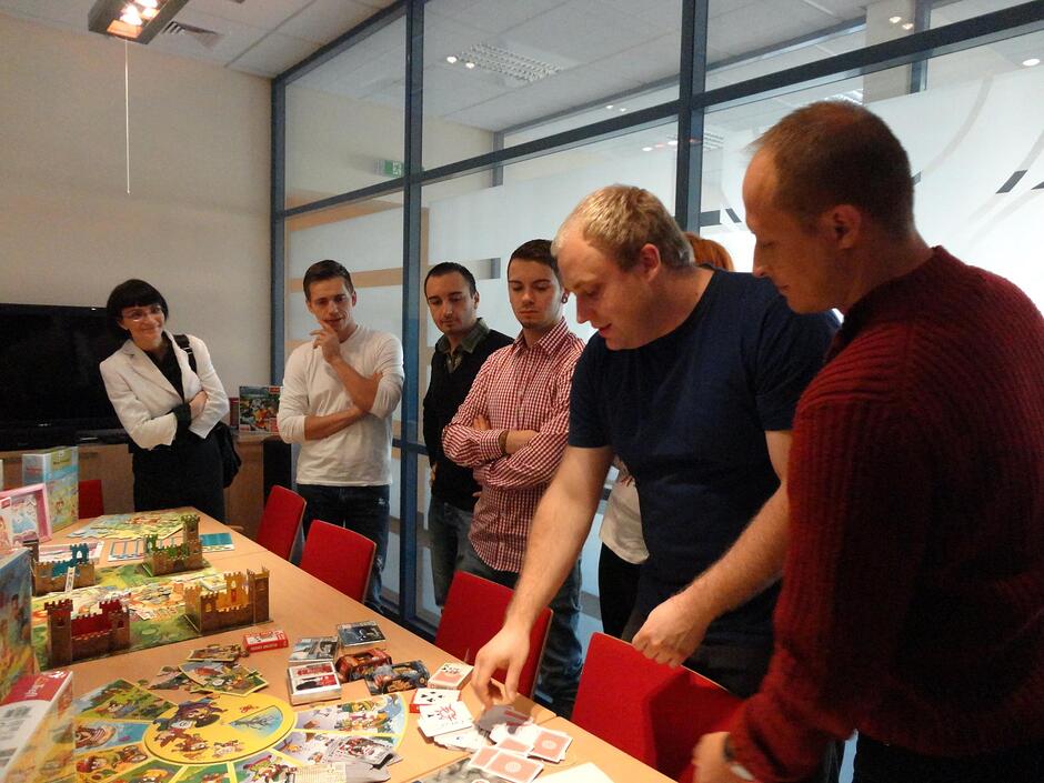 Grupa osób zgromadziła się w biurowym wnętrzu wokół stołu zastawionego różnorodnymi grami planszowymi i kartami. Dwaj mężczyźni na pierwszym planie z uwagą rozkładają elementy rozgrywki, podczas gdy pozostali uczestnicy z uśmiechem obserwują ich działania.