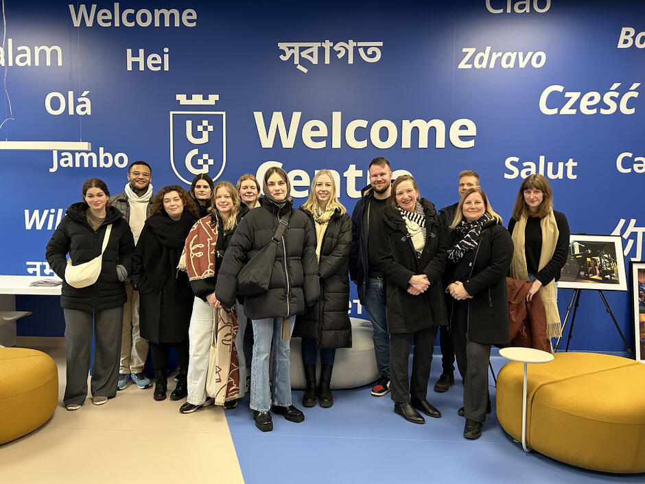 Grupa siedmiu osób pozuje do zdjęcia na zewnątrz, stojąc obok charakterystycznej, zielonej rzeźby przedstawiającej gdańskiego Neptuna. Uczestnicy spotkania są ubrani w stroje typu business casual i uśmiechają się na tle ceglanego, nowoczesnego budynku.