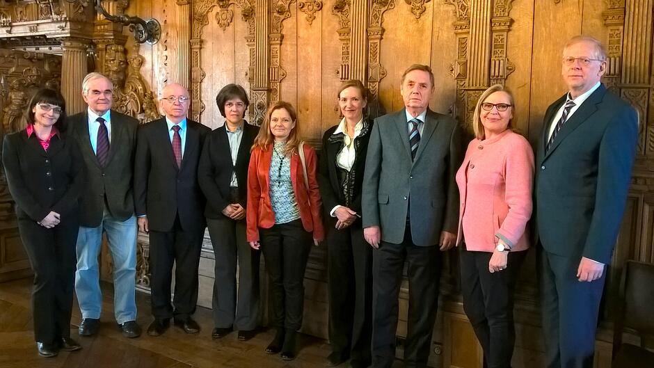 Na zdjęciu widać grupę dziewięciu osób w strojach wizytowych, pozujących do wspólnej fotografii w bogato zdobionym wnętrzu. Stoją oni na tle drewnianych, rzeźbionych paneli ściennych, które nadają pomieszczeniu zabytkowy charakter.
