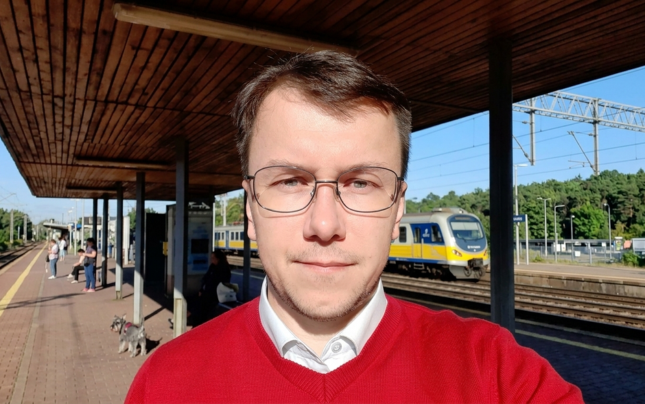 Na zdjęciu mężczyzna w czerwonym swetrze, w białej koszuli, w okularach, stojący na stacji kolejowej. W tle pociąg.