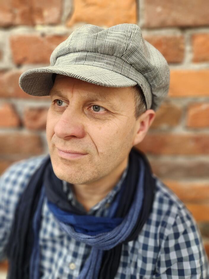 na zdjęciu mężczyzna w czapce z daszkiem, w kraciastej koszuli i przewieszonym na szyi szalikiem, w tle ściana z cegieł.