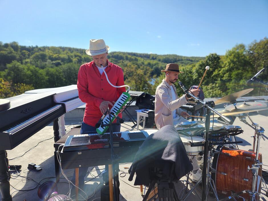 Dwóch muzyków występuje na świeżym powietrzu w słoneczny dzień. Za nimi rozciąga się zielony krajobraz z drzewami i jasnym, czystym niebem. Po lewej stronie stoi mężczyzna w czerwonej koszuli i jasnym kapeluszu. Gra na melodice, trzymając ją w dłoniach, a do ust prowadzi elastyczna rurka. Obok niego znajduje się czarno‑biały pianino ustawione na zewnątrz. Po prawej stronie drugi muzyk, ubrany w beżowy strój i brązowy kapelusz, siedzi przy zestawie perkusyjnym. Uderza w talerz perkusyjny miękką pałką. Wokół nich stoją mikrofony, statywy i kable, co sugeruje nagranie lub koncert plenerowy. Cała scena łączy naturalne otoczenie z atmosferą swobodnego, kreatywnego muzykowania.