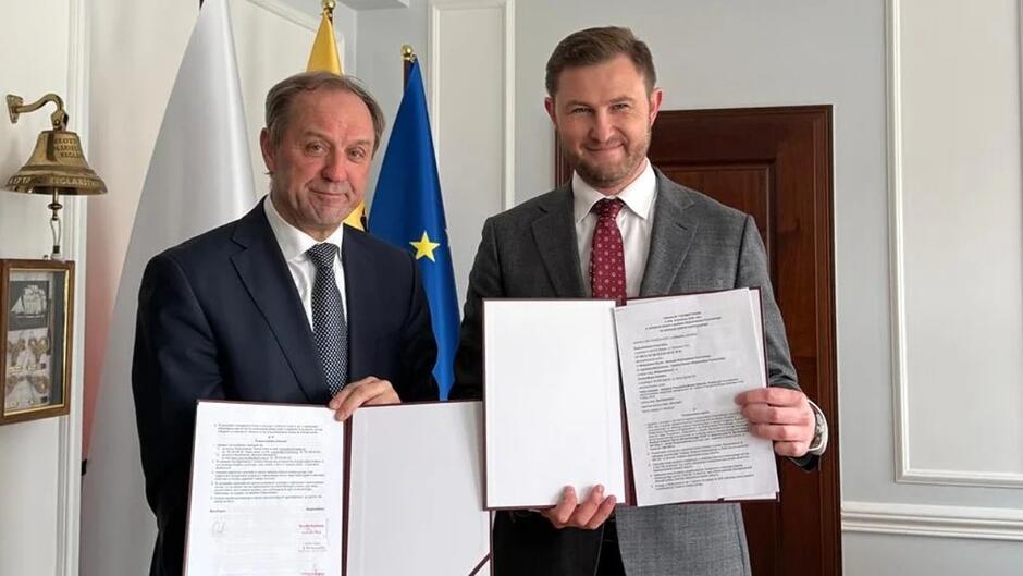 Dwaj mężczyźni w garniturach, uśmiechnięci, prezentują podpisane dokumenty