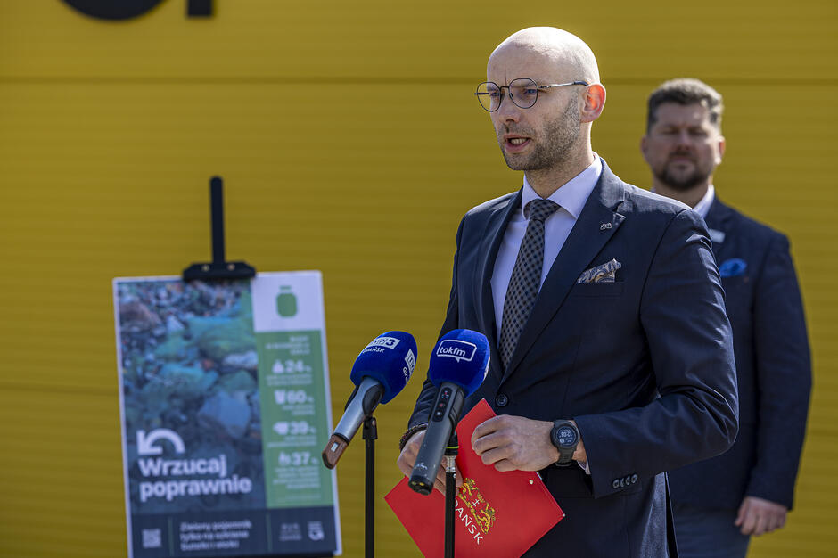 Na zdjęciu widać mężczyznę w okularach i garniturze, który przemawia do mikrofonów podczas oficjalnego spotkania lub konferencji prasowej. W tle znajduje się żółta ściana oraz tablica informacyjna dotycząca poprawnego segregowania odpadów.