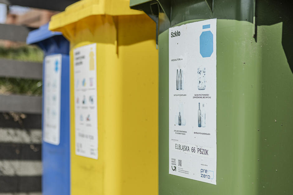 Na zdjęciu widać kolorowe pojemniki do segregacji odpadów z naklejonymi instrukcjami dotyczącymi wyrzucanych surowców. Zielony kosz na pierwszym planie jest przeznaczony na szkło, a obok niego stoją pojemniki żółty i niebieski.
