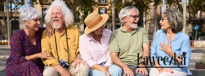 Zdjęcie przedstawia grupę pięciorga uśmiechniętych seniorów siedzących ramię w ramię na ławce w słoneczny dzień. Grupa składa się z trzech kobiet i dwóch mężczyzn, którzy wyglądają na zrelaksowanych i zaangażowanych we wspólną rozmowę.
Osoby: Seniorzy są ubrani w kolorowe, codzienne stroje – m.in. żółtą koszulę, kwiecistą sukienkę oraz błękitną bluzkę. Jeden z mężczyzn ma przewieszony przez ramię aparat fotograficzny, a jedna z kobiet nosi stylowy słomkowy kapelusz.
Emocje: Wszyscy na zdjęciu szczerze się śmieją lub uśmiechają, co sugeruje radosną, przyjacielską atmosferę.
Tło: W tle widać rozmytą architekturę miejską oraz drzewa, co wskazuje na przestrzeń publiczną, taką jak plac lub deptak.
Napis: W prawym dolnym rogu znajduje się czarny, ozdobny napis „Ławeczka”.
Całość emanuje pozytywną energią i celebruje aktywne życie towarzyskie seniorów.