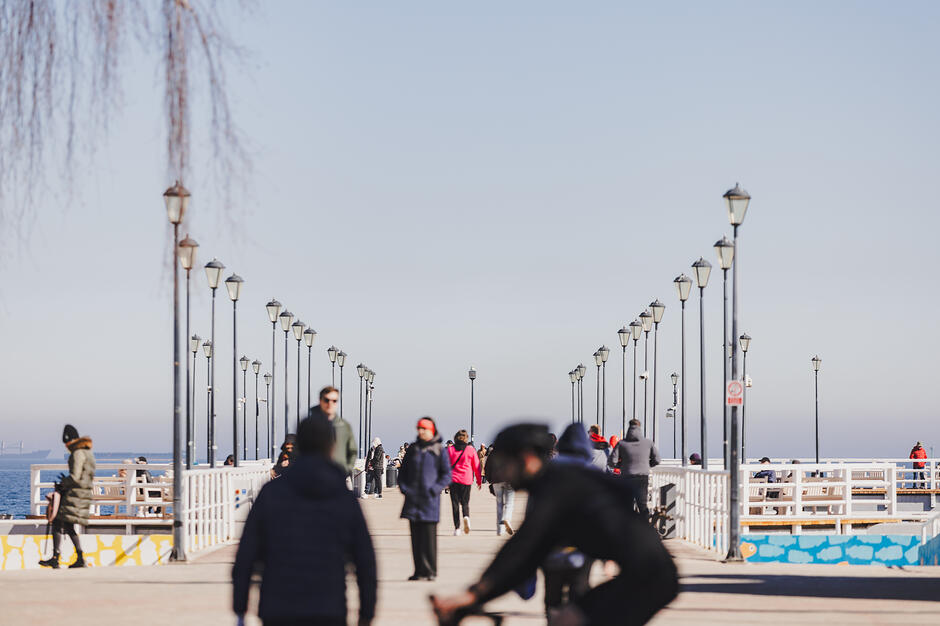 Zdjęcie przedstawia słoneczny dzień na molo w Gdyni Orłowie, po którym spacerują liczne osoby. Charakterystycznym elementem kompozycji są dwa rzędy stylowych latarni prowadzące wzrok w stronę morza.