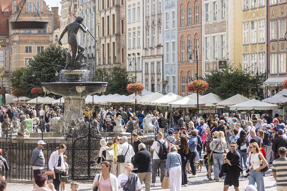 Zdjęcie przedstawia tętniący życiem Długi Targ w Gdańsku z centralnie umieszczoną Fontanną Neptuna, wokół której gromadzą się liczni turyści. W tle widać rzędy kolorowych, zabytkowych kamienic oraz liczne kawiarniane ogródki pod białymi parasolami.