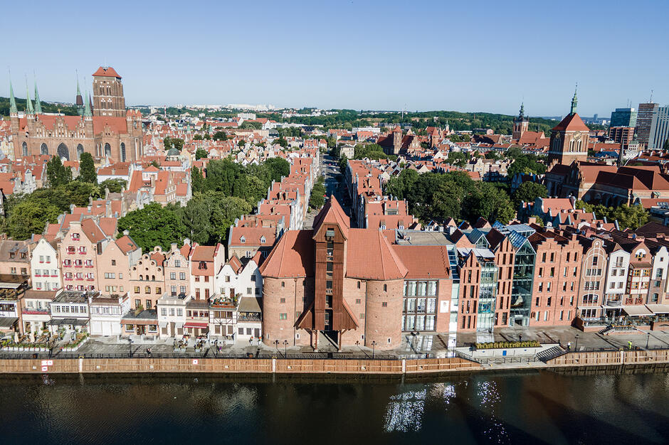 Zdjęcie przedstawia panoramę gdańskiego nabrzeża Motławy z charakterystycznym Żurawiem i rzędami zabytkowych kamienic o czerwonych dachach. W tle góruje potężna Bazylika Mariacka, która dominuje nad gęstą zabudową Starego Miasta.
