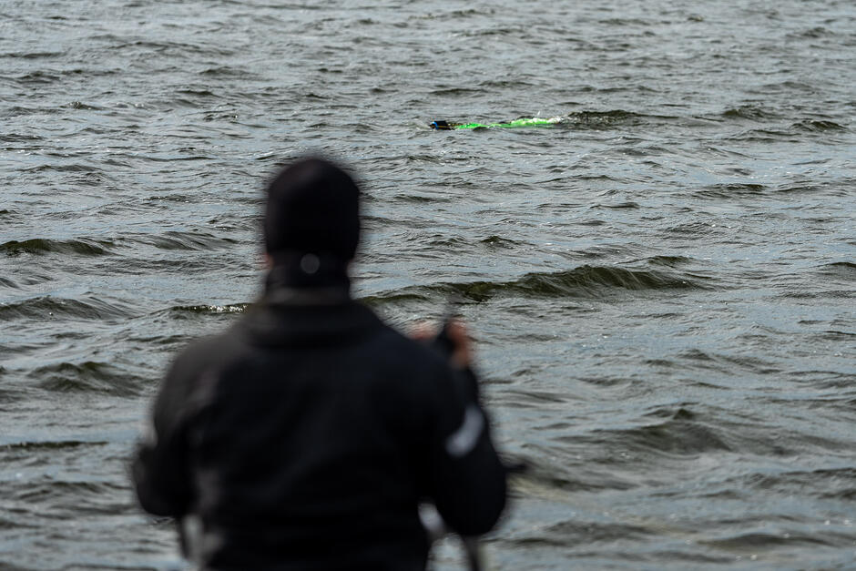 Na pierwszym planie widać osobę w ciemnej odzieży, która obserwuje lub zdalnie steruje obiektem znajdującym się na tafli wody. W oddali pośród fal przemieszcza się jaskrawozielone, podłużne urządzenie przypominające drona podwodnego.