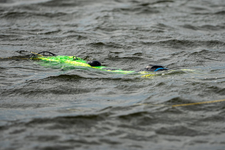 Jaskrawozielone, podłużne urządzenie podwodne częściowo zanurzone w ciemnej, pofalowanej wodzie przemieszcza się po jej powierzchni. Obiekt posiada czarne elementy konstrukcyjne i wydaje się być zdalnie sterowanym dronem lub sonarem przeznaczonym do badań wodnych.