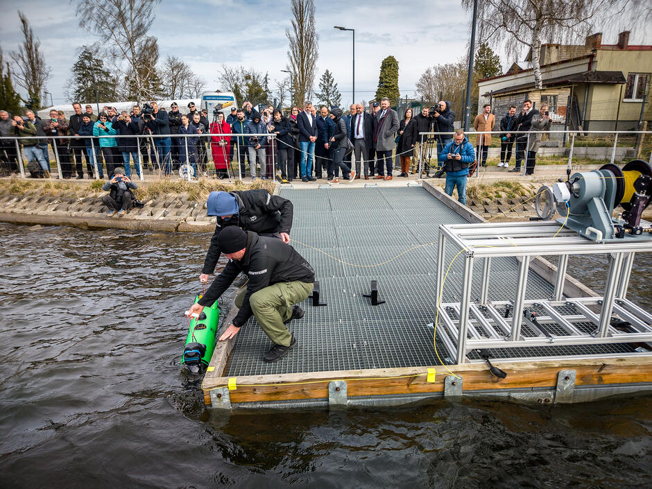Zdjęcie przedstawia dwóch mężczyzn na pływającym pomoście, którzy opuszczają do wody zielone urządzenie podwodne, prawdopodobnie drona lub sonar. Całemu wydarzeniu z brzegu przygląda się liczna grupa osób, w tym dziennikarze i oficjele, co sugeruje oficjalną prezentację lub testy technologiczne.