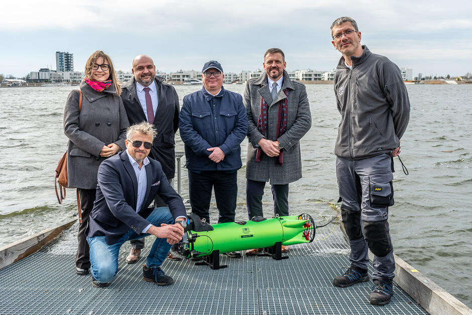 Na pomoście stoi grupa sześciu osób pozujących do zdjęcia z nowoczesnym, zielonym urządzeniem podwodnym. W tle widać zbiornik wodny oraz zabudowania miejskie pod zachmurzonym niebem.