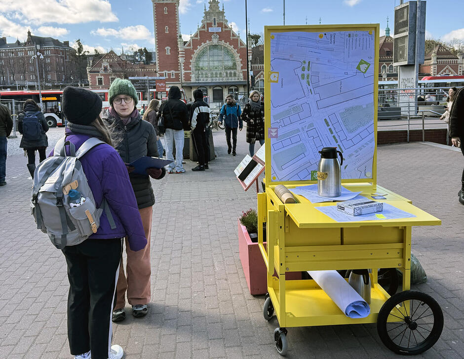 Na miejskim placu stoi mobilne stanowisko konsultacji, obok kobieta tłumaczy coś drugiej kobiecie.