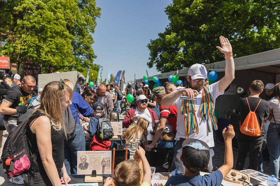 Tłum ludzi w ładną, słoneczna pogodę bierze udział w plenerowym wydarzeniu. Na pierwszym planie mężczyzna w czapeczce przeprowadza eksperyment naukowy, obserwują go dzieci.