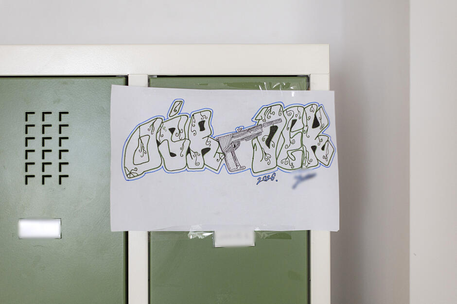 Zdjęcie przedstawia rysunek w stylu graffiti wykonany na białej kartce, która jest przyklejona do metalowych, zielonych szafek ubraniowych. Napis charakteryzuje się grubymi literami z niebieskim obrysem i detalami przypominającymi pistolet maszynowy oraz ozdobne wzory.