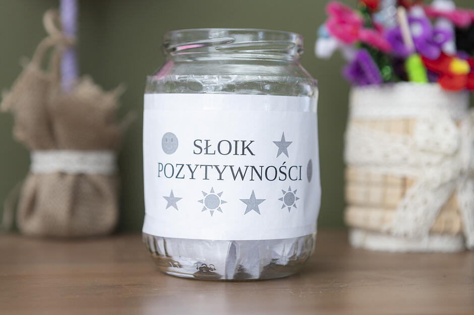 Na środku zdjęcia znajduje się szklany słoik z naklejoną białą etykietą, na której widnieje napis „SŁOIK POZYTYWNOŚCI” ozdobiony motywami gwiazd i słońc. Wewnątrz naczynia widać zwinięte karteczki, a w tle rozmyte elementy dekoracyjne, które tworzą przytulną atmosferę.