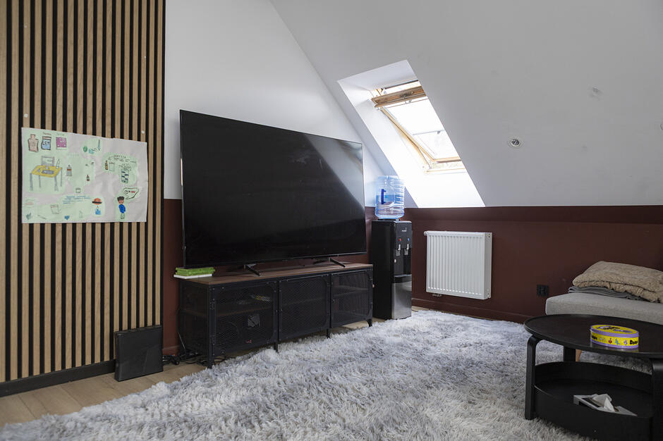 Zdjęcie przedstawia nowocześnie urządzone poddasze z dużym telewizorem umieszczonym na niskiej, metalowej szafce. Wnętrze charakteryzuje się skośnym sufitem z oknem połaciowym, puszystym szarym dywanem oraz dekoracyjnymi lamelami ściennymi, na których wisi dziecięcy rysunek.