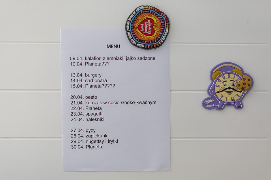 Zdjęcie przedstawia kartkę z jadłospisem na kwiecień, przypiętą magnesami do białej powierzchni, prawdopodobnie lodówki. W menu widnieją konkretne potrawy, takie jak carbonara czy naleśniki, przeplatane tajemniczymi wpisami „Planeta”, którym towarzyszą znaki zapytania.