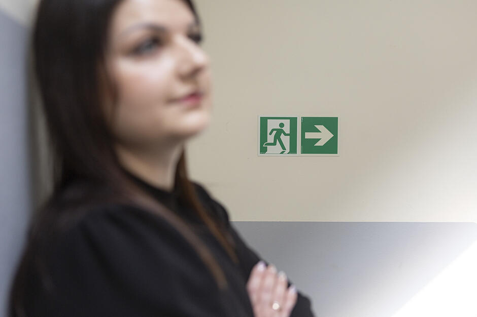 Na zdjęciu widać nieostrą sylwetkę kobiety o ciemnych włosach, stojącej z założonymi rękami na tle jasnej ściany. Głównym, wyraźnym punktem kompozycji jest umieszczony po prawej stronie zielony znak wyjścia ewakuacyjnego z białą strzałką.