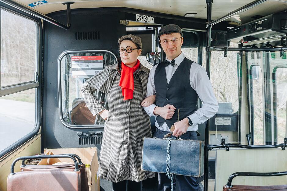 Dwie osoby patrzą prosto w obiektyw. Stoją w zabytkowym autobusie. Są przeprani w ubrania z okresu Międzywojnia 